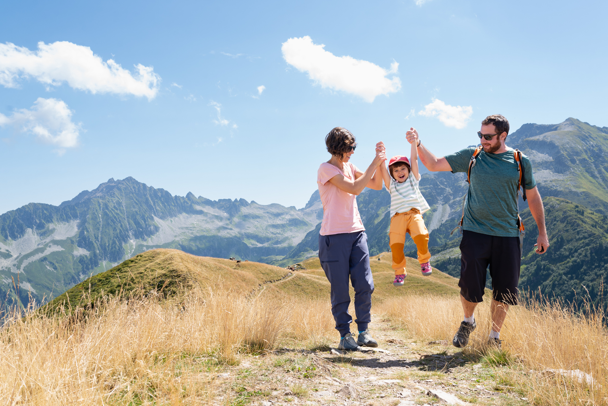 Mother, father holding hands of their little daughter and helping her to jump during a hiking day in the French alps at summer season.